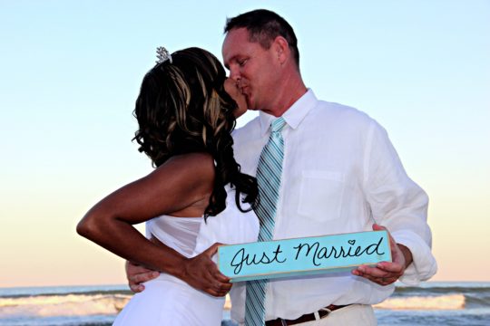bride and groom kissing on the beach holding just married sign