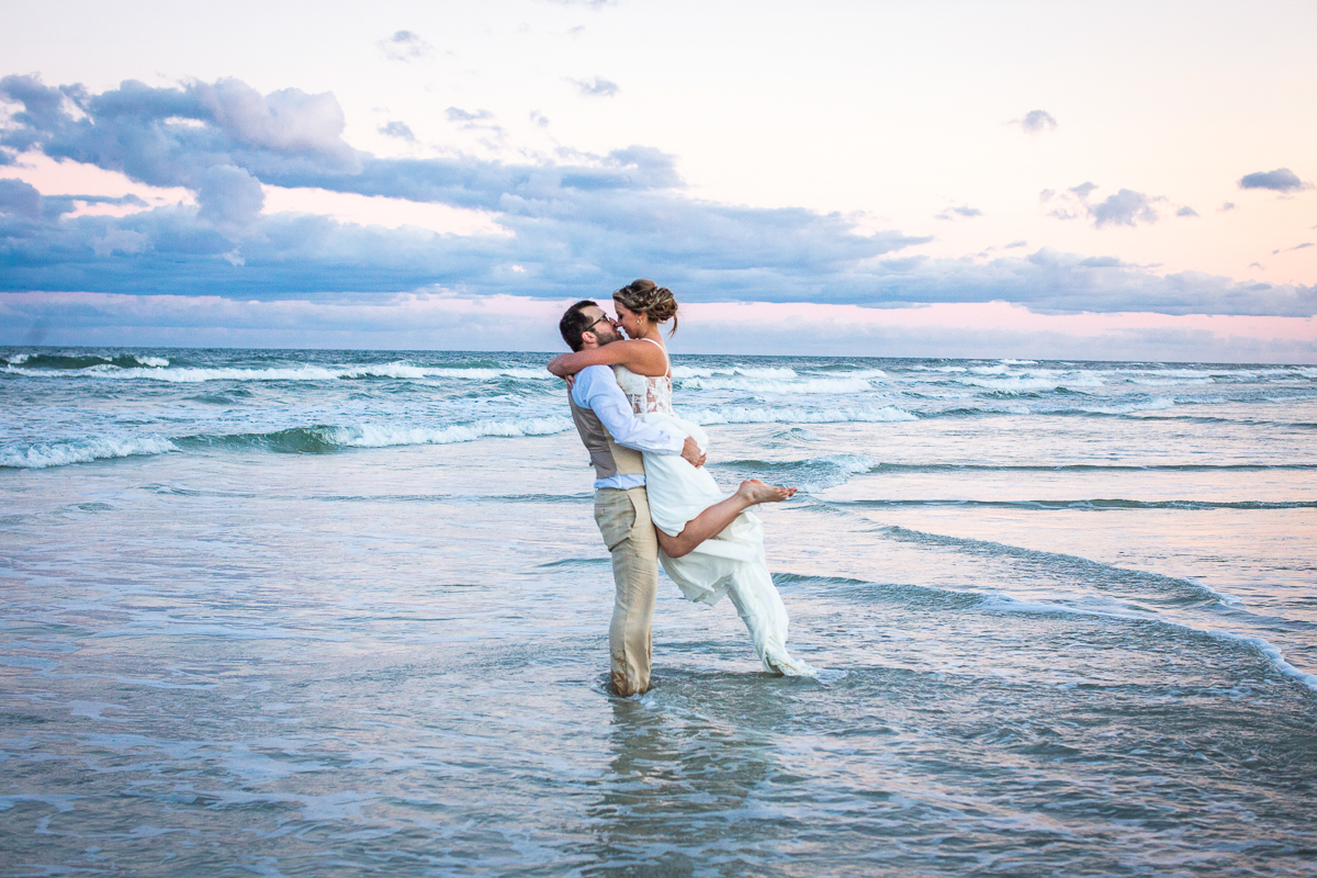 groom holding bride as they stand in the water at the beach and the sunset is behind them